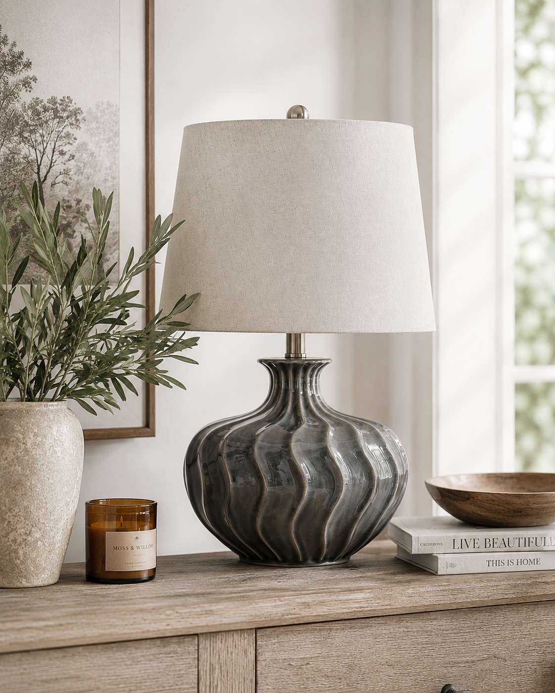 A table lamp with a ribbed, textured dark gray ceramic base and a beige fabric drum shade, styled on a light wood console table in a bright, modern room.